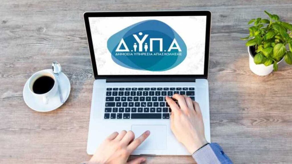 Ποιοι άνεργοι δικαιούνται έξτρα επίδομα 270 ευρώ το μήνα 1 Κάτοψη: Ανοικτό λαπτοπο πάνω σε γραφείο με δύο χέρια που πληκτρολογούν και στην οθόνη λογότυπο ΔΥΠΑ