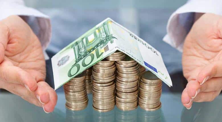 ένα σπιτάκι από νομίσματα και η οροφή του έχει δημιουργηθεί από χαρτονομίσματα δύο χέρια πλησιάζουν να το στηρίξουν